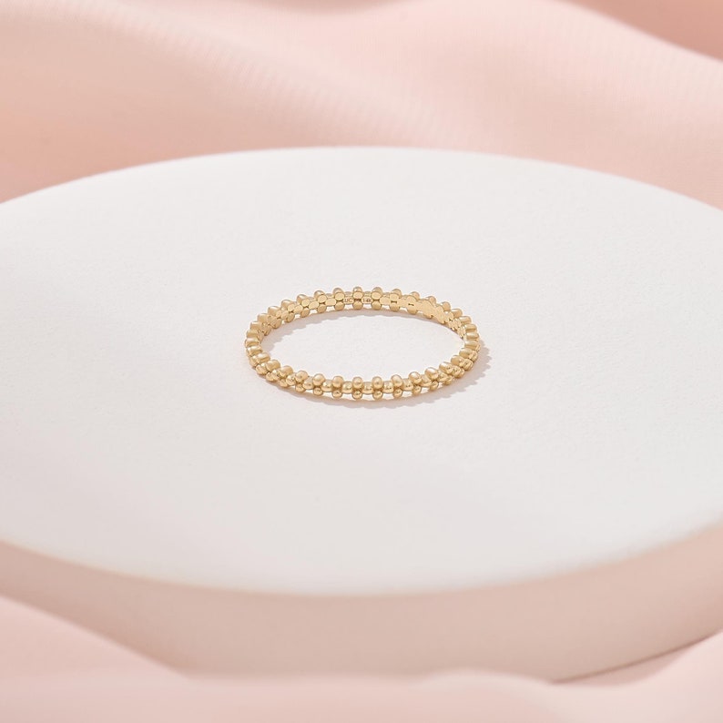 Op de afbeelding: Een dunne gouden ring met een doorlopende band van kleine, uniforme bolvormige kralen gerangschikt in een delicaat, cirkelvormig patroon. Het metaal heeft een gepolijste afwerking die het licht reflecteert op het oppervlak van elke kraal.