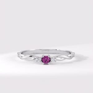 Pode incluir: Um anel de tom prateado com uma pedra preciosa magenta de corte redondo ao centro, ladeada por duas pedras claras mais pequenas. A banda apresenta um design torcido, semelhante a uma corda, em ambos os lados, criando um visual elegante.