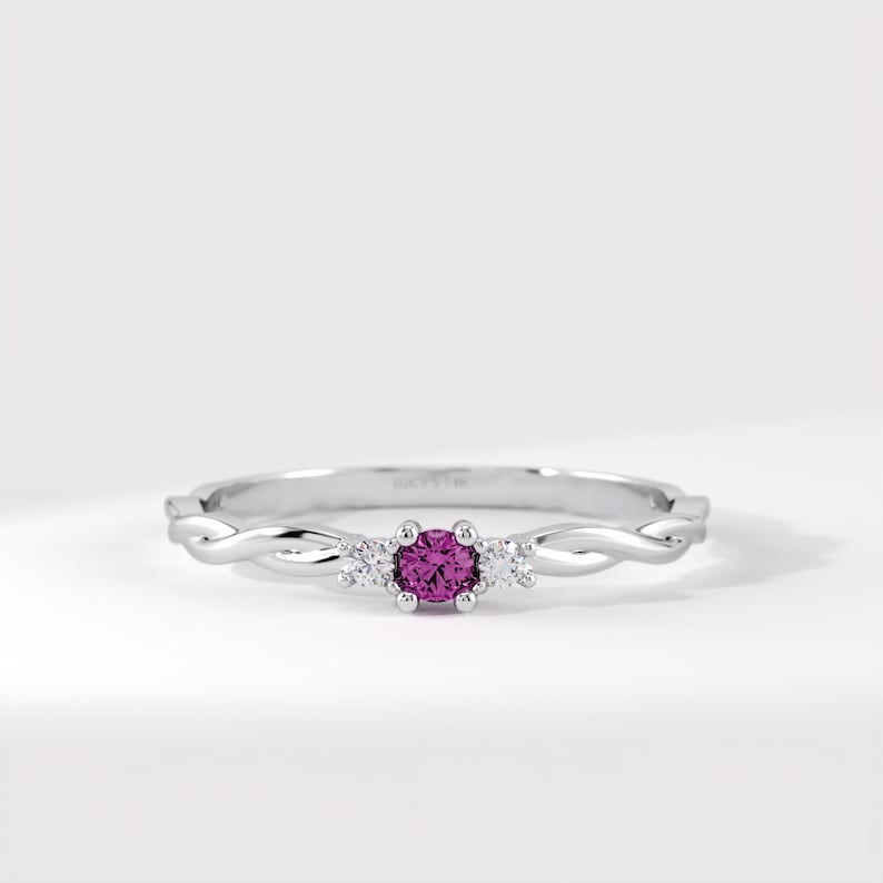 Pode incluir: Um anel de tom prateado com uma pedra preciosa magenta de corte redondo ao centro, ladeada por duas pedras claras mais pequenas. A banda apresenta um design torcido, semelhante a uma corda, em ambos os lados, criando um visual elegante.