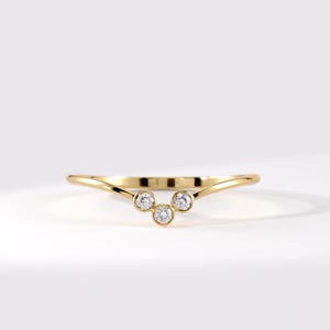 May include: A gold V-shaped ring featuring three round, bezel-set clear stones arranged in a cluster at the center point. The band has a polished, smooth finish and a curved silhouette that tapers toward the sides, creating a delicate appearance.