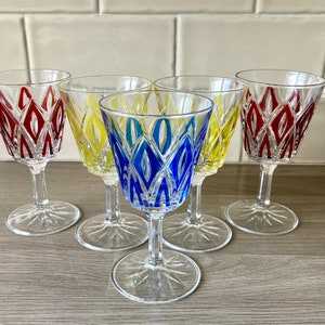 May include: Six clear crystal wine glasses with colourful geometric designs. The glasses feature red, yellow, and blue patterns. Each glass has a clear stem and a starburst base, ideal for serving wine or other drinks.