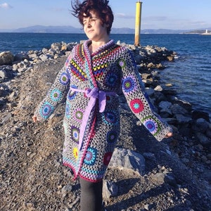 Op de afbeelding: Een grijze, handgemaakte gehaakte omslagdoek met kleurrijke bloemen- en geometrische patronen. De omslagdoek heeft een paarse riem en wordt buiten bij de zee gedragen. Het haakontwerp bevat tinten roze, blauw, groen en geel.