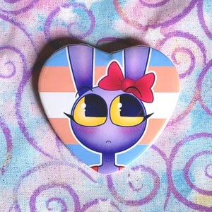 May include: A heart-shaped pin featuring a purple cartoon rabbit character with large yellow eyes and a red bow on its head. The background of the pin displays horizontal stripes in light blue, pink, and white, creating a vibrant and colorful design.