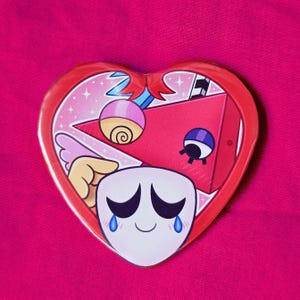 May include: A heart-shaped pin with a red border and a cartoon design. The design features a white face with black eyes and blue tears, a red triangle, and other colorful elements. The background is pink.