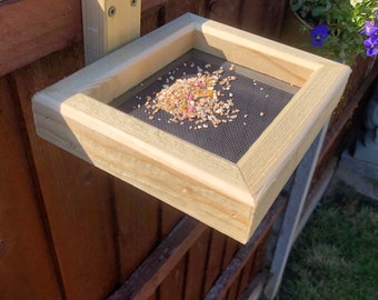 Op de afbeelding: Een houten vogelvoederhuisje met een gaasbodem om te voorkomen dat eekhoorns bij het vogelzaad kunnen. De voederbak is bevestigd aan een houten schutting.