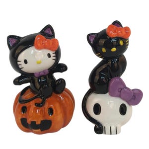 May include: Two ceramic Halloween figurines. One is a black cat dressed as Hello Kitty sitting on an orange pumpkin. The other is a black cat with an orange bow sitting on a white skull with a purple bow. Both have black ears and yellow eyes.