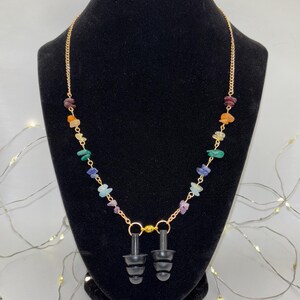 May include: A gold-colored chain necklace with colorful gemstone beads and two black earplugs. The necklace is displayed on a black mannequin. The gemstones are various colors, including red, orange, yellow, green, blue, and purple.