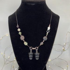 May include: A black necklace with green, purple, and white beads, a lava rock bead, and a silver charm. The necklace has black earplugs attached to it.