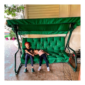 May include: Two children sitting on a green cushioned swing set with a green canopy. The swing set is on a patio with a brick floor.