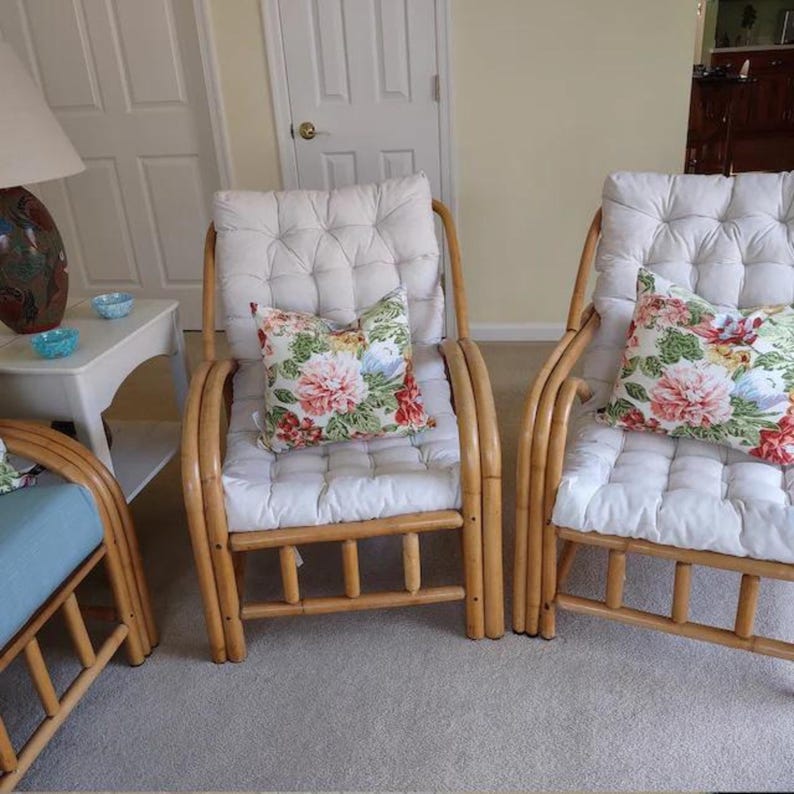 Peut inclure: Deux chaises en osier &agrave; coussin blanc avec des coussins &agrave; motifs floraux. Les chaises sont dans un salon avec une table d'appoint blanche et une porte blanche en arri&egrave;re-plan.