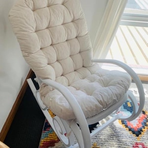 Peut inclure: Fauteuil &agrave; bascule blanc avec un coussin rembourr&eacute;. Le coussin est capitonn&eacute; et fait de tissu beige clair. Le fauteuil est en bois et a une finition blanche.