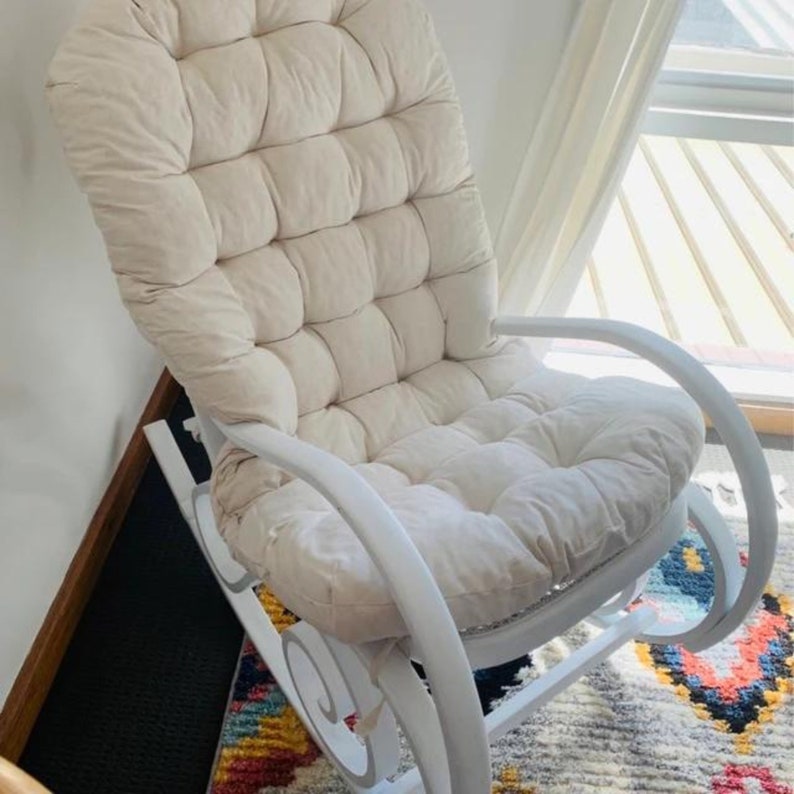 Peut inclure: Fauteuil &agrave; bascule blanc avec un coussin rembourr&eacute;. Le coussin est capitonn&eacute; et fait de tissu beige clair. Le fauteuil est en bois et a une finition blanche.