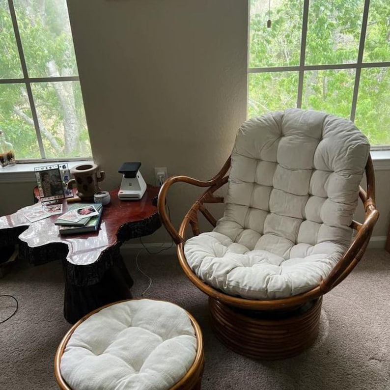 Peut inclure: Un fauteuil pivotant en osier brun avec un coussin blanc et un pouf blanc assorti. Le fauteuil et le pouf sont dans un salon avec une table en bois et une fen&ecirc;tre en arri&egrave;re-plan.