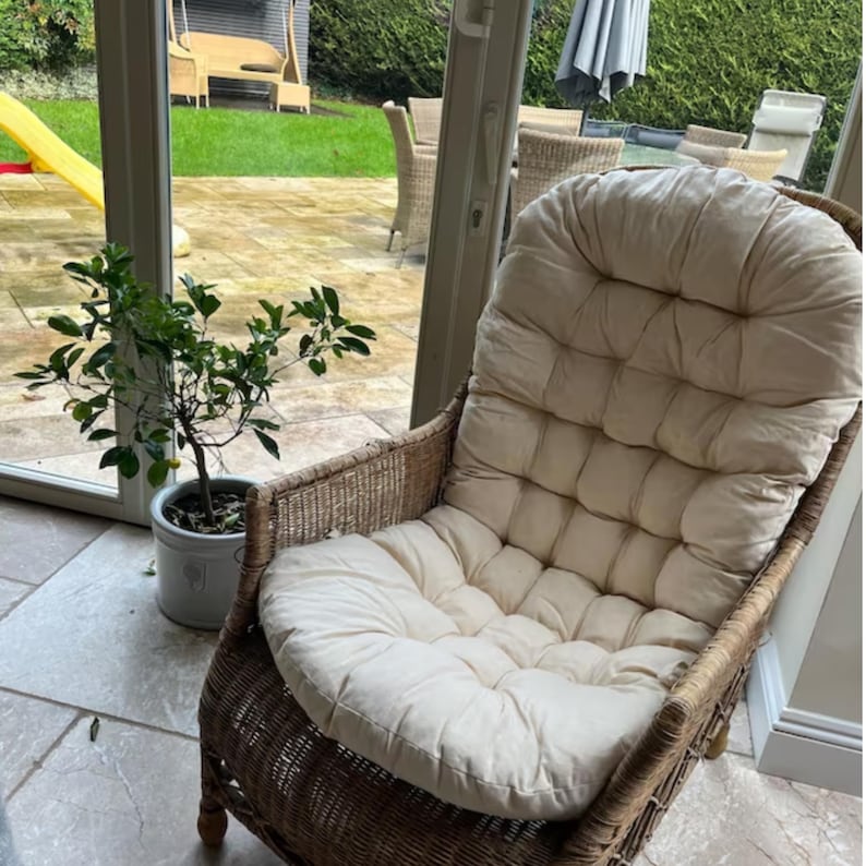 Peut inclure: Un fauteuil en osier avec un coussin blanc. Le coussin a un design capitonn&eacute; et est tr&egrave;s confortable. Le fauteuil est dans une pi&egrave;ce avec une fen&ecirc;tre et une plante.