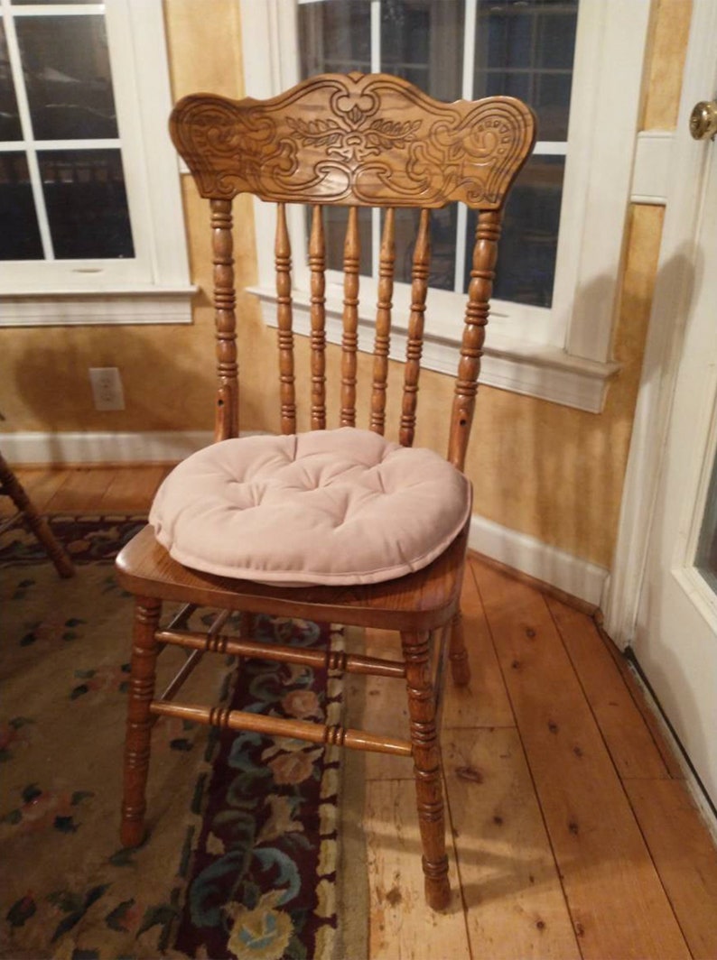 May include: A wooden chair with a carved back and a light pink cushion. The chair is sitting on a patterned rug in front of a window.