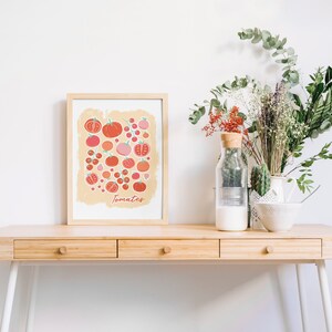 Pu&ograve; includere: Una stampa incorniciata di un'illustrazione colorata di pomodori su uno sfondo beige chiaro. La stampa &egrave; intitolata "Tomatoes" ed &egrave; esposta su una scrivania di legno con un vaso di fiori secchi e una ciotola di ceramica bianca.