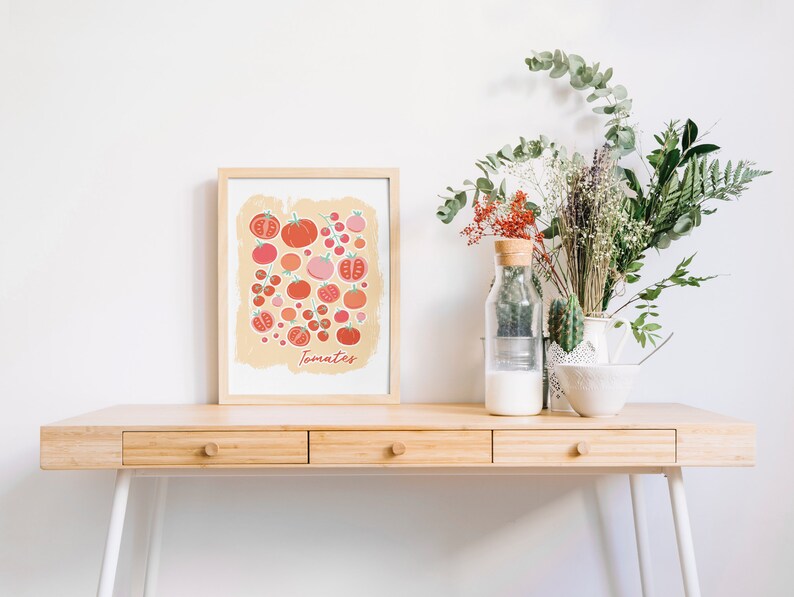 Pu&ograve; includere: Una stampa incorniciata di un'illustrazione colorata di pomodori su uno sfondo beige chiaro. La stampa &egrave; intitolata "Tomatoes" ed &egrave; esposta su una scrivania di legno con un vaso di fiori secchi e una ciotola di ceramica bianca.