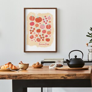 Pu&ograve; includere: Una stampa incorniciata di un'illustrazione colorata di pomodori su uno sfondo beige. La stampa ha la parola "Tomates" scritta in basso. La stampa &egrave; appesa a un muro bianco sopra un tavolo di legno con una teiera, una ciotola di noci, una ciotola di croissant e un barattolo di vetro.