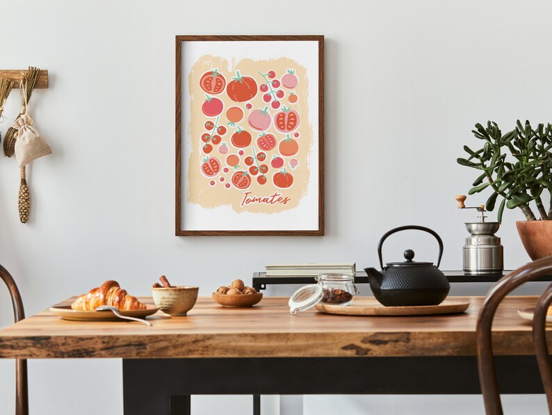 Pu&ograve; includere: Una stampa incorniciata di un'illustrazione colorata di pomodori su uno sfondo beige. La stampa ha la parola "Tomates" scritta in basso. La stampa &egrave; appesa a un muro bianco sopra un tavolo di legno con una teiera, una ciotola di noci, una ciotola di croissant e un barattolo di vetro.