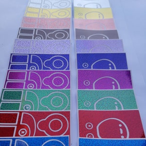 以下が含まれることがあります： シンプルなデザインの、キラキラ光るカラフルなステッカー16枚セット。各ステッカーには、2つの目と口のある顔の白いアウトラインが描かれています。ステッカーは縦に積み重ねられており、色はシルバーからブルーまで変化しています。