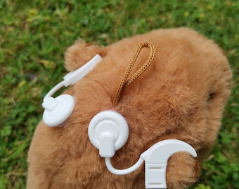 May include: A brown plush capybara toy featuring a white cochlear implant accessory attached to the side of its head. The accessory includes a white sound processor, a circular coil, and a thin white connecting wire, with a small brown loop at the top.