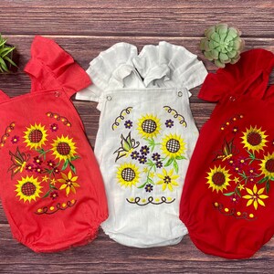 May include: Three baby onesies with sunflower embroidery. One onesie is red, one is white, and one is a light pink. All three onesies have ruffled sleeves.