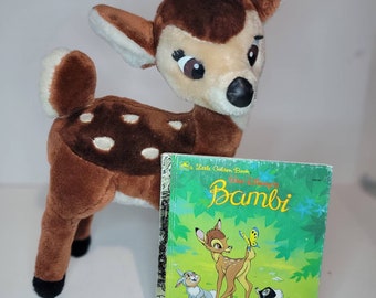 Puede incluir: Un juguete de peluche Bambi de color marrón con manchas blancas y pezuñas negras. El juguete está de pie junto a un pequeño libro dorado titulado "Walt Disney's Bambi".