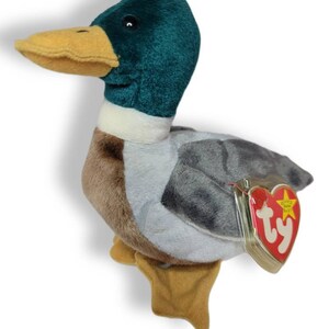 May include: A green, brown, and gray plush duck with a yellow beak and a Ty Beanie Baby tag. The tag is red with a white star and the word "Ty" in white.