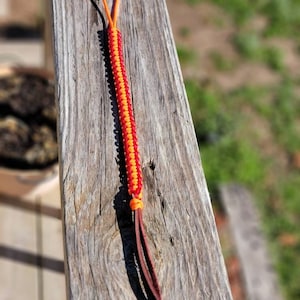 May include: A handmade lanyard with a braided orange and red design. The lanyard has a loop at the top and a leather tassel at the bottom. The lanyard is resting on a weathered wooden surface.