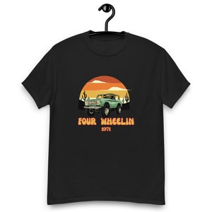 Puede incluir: Una camiseta negra con un gráfico de estilo vintage de un Ford Bronco verde de 1971 conduciendo por un paisaje desértico con el texto "Four Wheelin' 1971".