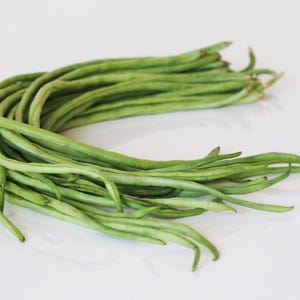 May include: A bundle of fresh, vibrant green long beans, also known as yardlong beans, arranged in a curved shape. The beans are smooth and slender, with a slight curve, and are set against a plain white background.