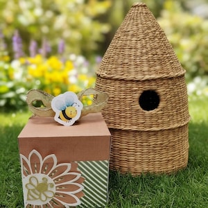 May include: A woven straw beehive with a circular entrance hole sits next to a square box. The box features a light brown lid, a green and white striped side, and a white flower cutout with a gold glitter bee and a small bee sticker on top.