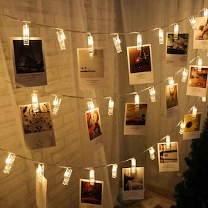 May include: A string of white fairy lights with clear plastic clips holding up photos. The lights are strung across a wall and are turned on, casting a warm glow.