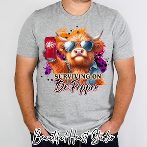 May include: A grey t-shirt featuring a cartoon Highland cow wearing sunglasses and holding a can of Dr Pepper. The text "SURVIVING ON Dr. Pepper" is printed on the shirt.
