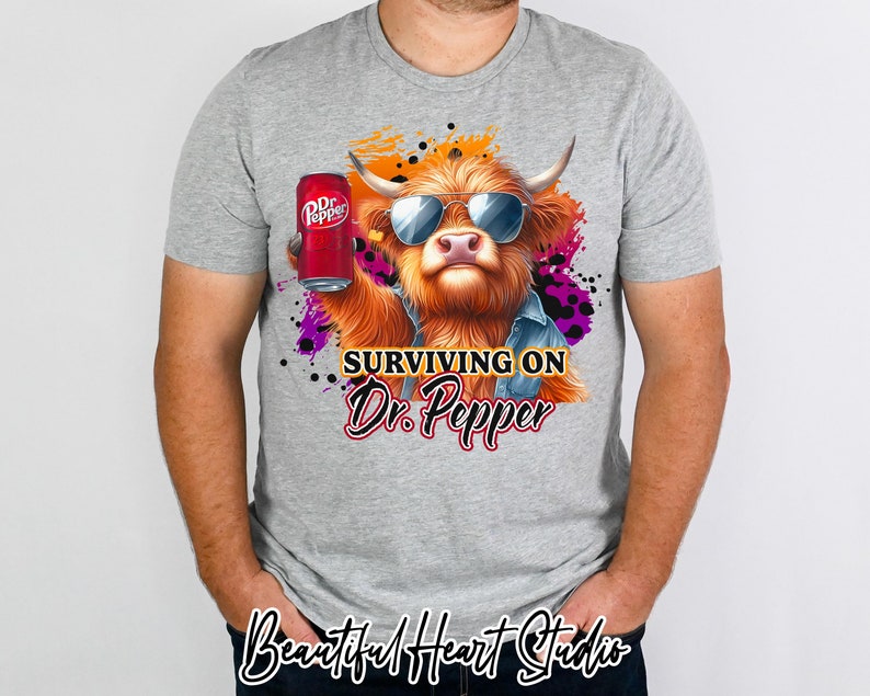 May include: A grey t-shirt featuring a cartoon Highland cow wearing sunglasses and holding a can of Dr Pepper. The text "SURVIVING ON Dr. Pepper" is printed on the shirt.