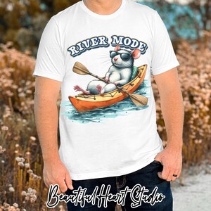 K&ouml;nnte beinhalten: Wei&szlig;es T-Shirt mit einer Cartoon-Ratte mit Sonnenbrille, die ein Kajak paddelt. Der Text "RIVER MODE" befindet sich &uuml;ber der Ratte. Das Bild ist im Aquarellstil mit Blau-, Orange- und Braunt&ouml;nen gehalten.