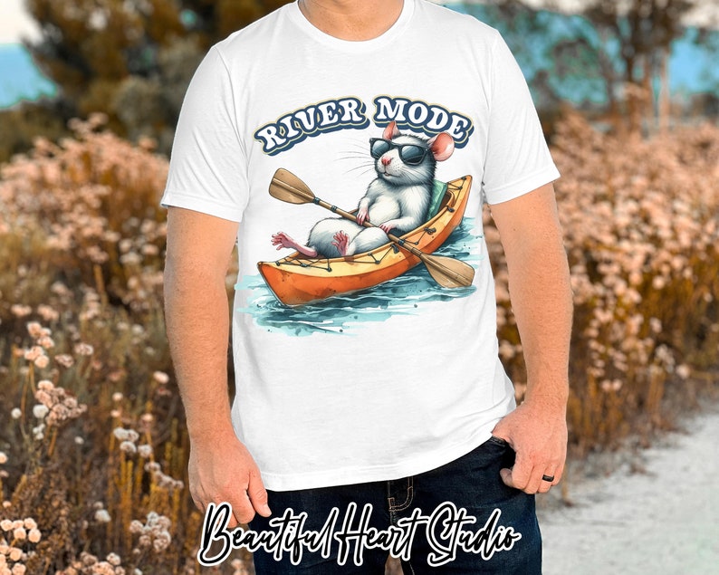 K&ouml;nnte beinhalten: Wei&szlig;es T-Shirt mit einer Cartoon-Ratte mit Sonnenbrille, die ein Kajak paddelt. Der Text "RIVER MODE" befindet sich &uuml;ber der Ratte. Das Bild ist im Aquarellstil mit Blau-, Orange- und Braunt&ouml;nen gehalten.