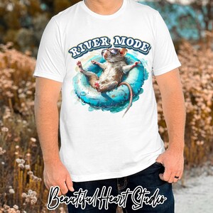 Puede incluir: Camiseta blanca con un gr&aacute;fico de una rata con gafas de sol flotando en un flotador inflable azul. El texto "River Mode" est&aacute; impreso en amarillo sobre la rata.