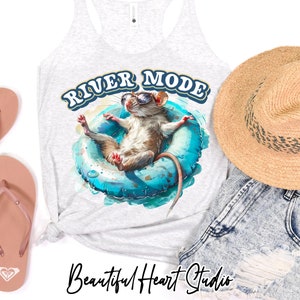Puede incluir: Una camiseta sin mangas gris con el texto "River Mode" en letras azules y blancas. Un rat&oacute;n de dibujos animados con gafas de sol flota en un flotador inflable azul. El rat&oacute;n es marr&oacute;n con la barriga blanca. La camiseta sin mangas est&aacute; sobre un fondo blanco con un par de chanclas rosas y un sombrero de paja.