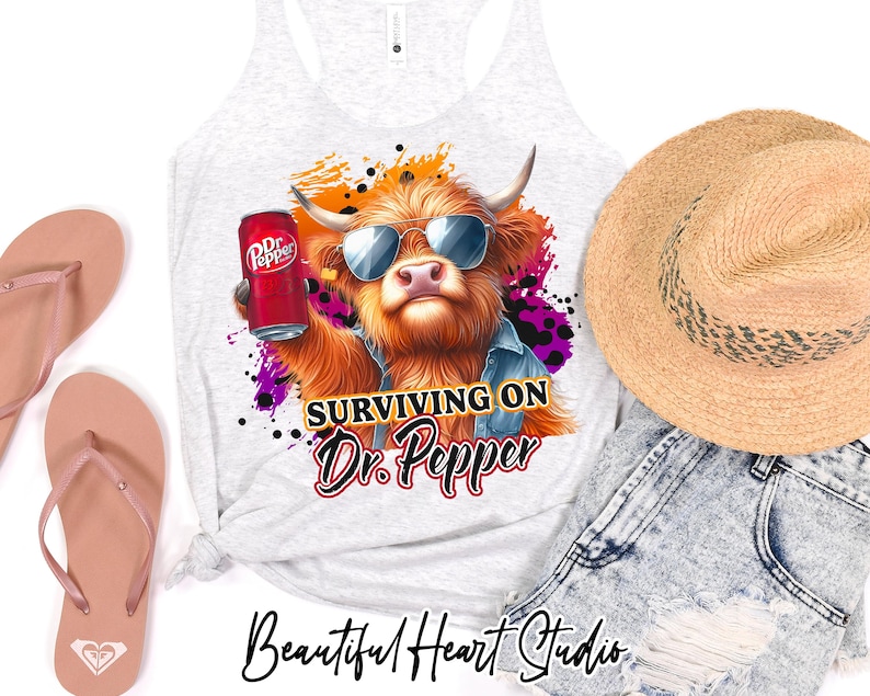 May include: A white tank top featuring a graphic of a Highland cow wearing sunglasses and holding a can of Dr Pepper. The text "Surviving on Dr. Pepper" is beneath the cow. The tank top is displayed against a white backdrop.