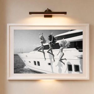 Könnte beinhalten: Schwarz-Weiß-Druck mit drei Figuren, die von einer Yacht springen. Das Bild ist an einer beigefarbenen Wand angebracht, mit einer dunklen Bronze-Leuchte darüber. Der Raum hat einen hellen Holzboden.