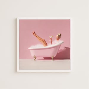 May include: A framed print featuring a woman relaxing in a pink clawfoot bathtub filled with bubbles. She holds a cocktail and has her legs raised. The background is a solid pink color.
