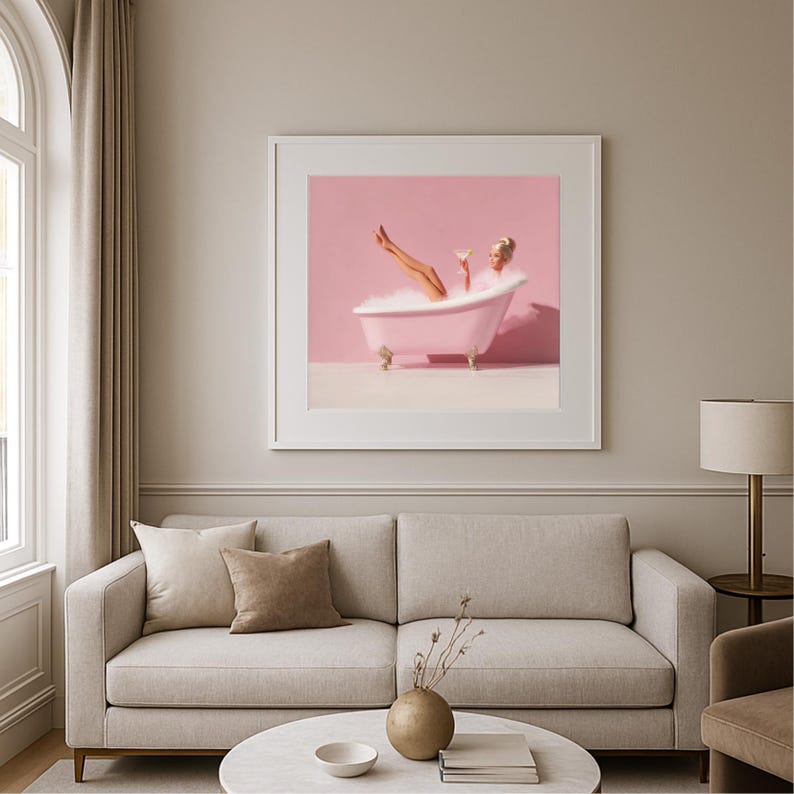 May include: A framed print depicts a woman in a pink bathtub with bubbles, holding a cocktail. The artwork is set against a pink backdrop, with the tub resting on a white surface. The print is displayed above a beige sofa in a well-lit room.