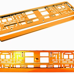 May include: Two orange-colored car license plate frames, one assembled and one disassembled, are displayed against a white background. The frames have a shiny, metallic finish and a rectangular shape with various cutouts and mounting holes.