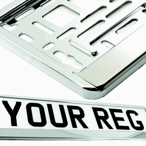 May include: A chrome license plate frame and a license plate with the text "YOUR REG" in black. The frame is rectangular with a shiny, reflective surface. The license plate is white with black lettering.