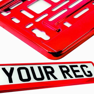 May include: A red license plate frame and plate. The frame is a shiny, metallic red with cut-out sections. The plate is white with black text that reads "YOUR REG" in a bold font. The items are against a white background.