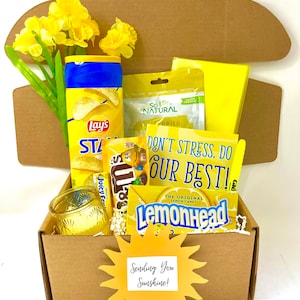 May include: A yellow gift box filled with snacks and a yellow sun cutout with the text "Sending You Sunshine!"  The box contains a bag of Lay's potato chips, a bag of Lemonhead sweets, a bag of Sour Patch Kids sweets, a bag of dried fruit, a yellow candle, and a yellow flower.