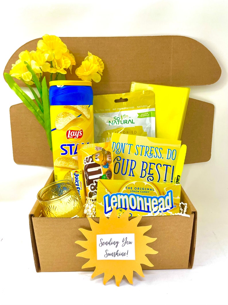 May include: A yellow gift box filled with snacks and a yellow sun cutout with the text "Sending You Sunshine!"  The box contains a bag of Lay's potato chips, a bag of Lemonhead sweets, a bag of Sour Patch Kids sweets, a bag of dried fruit, a yellow candle, and a yellow flower.
