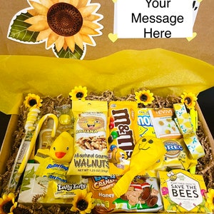 May include: A yellow gift box filled with various yellow-themed snacks and treats. The box includes a yellow candy cane, a yellow rubber duck, a bag of walnuts, a bag of M&Ms, a bag of lemon-flavoured sweets, a bag of graham crackers, a bag of Laffy Taffy, a bag of Golden Oreo biscuits, a bag of sunflower seeds, and a small bag of wildflower seeds. The box is decorated with a yellow sunflower and the words "Your Message Here".