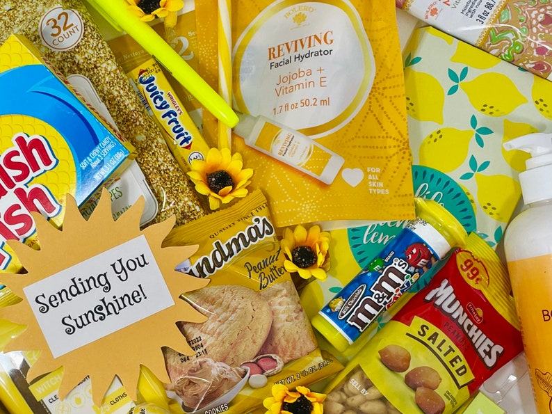 May include: A yellow gift basket filled with various items, including snacks, sweets, and beauty products. The basket includes a sun-shaped card that says "Sending You Sunshine!" and a variety of yellow and gold items, such as a yellow pencil, a yellow and gold bag of peanut butter biscuits, and a yellow and gold bag of M&Ms.